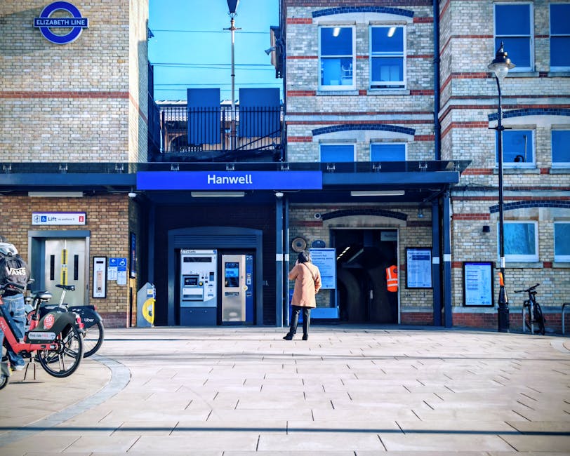 A woman standing in front of Hanwell station, part of the Elizabeth Line, with a modern brick building featuring large windows and a black canopy above the entrance. Several bicycles are parked on the